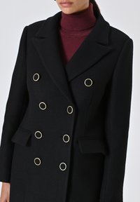 Cappotto doppiopetto nero con grandi bottoni circolari dorati, tessuto testurizzato, revers a punta e tasche laterali; indossato sopra un dolcevita bordeaux.