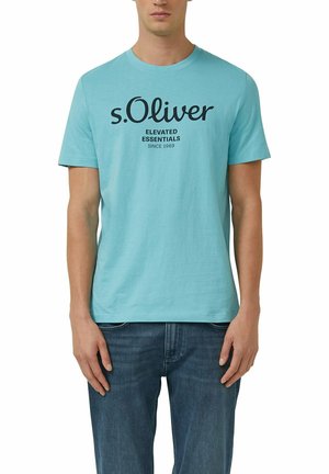 Hombre con camiseta de manga corta azul claro con el texto "s.Oliver Elevated Essentials Since 1969", combinado con jeans azules, de pie derecho.