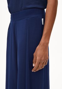 Navyblauwe wijde broek gemaakt van zachte stof, met een elastische tailleband en een klein merkembleem aan de zijkant. Glad textuur.