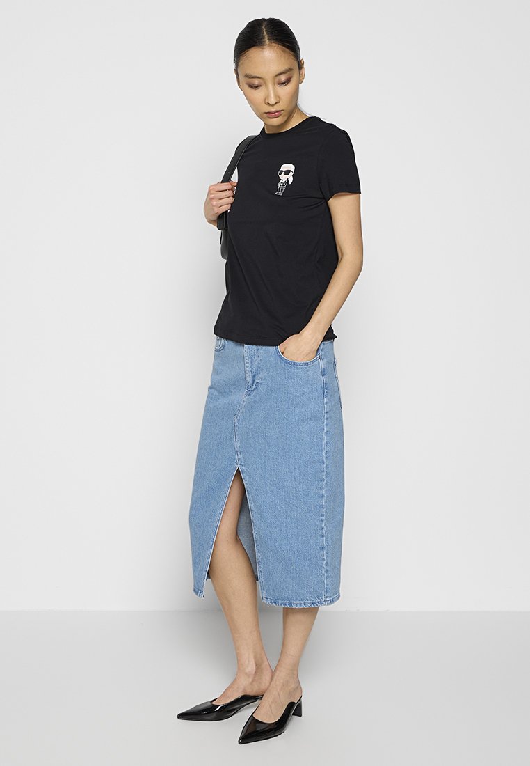 Schwarzes Baumwoll-T-Shirt mit einem kleinen Grafikdesign auf der Brust, kombiniert mit einem Jeansrock mit einem Schlitz vorne und schwarzen spitzen Schuhen.