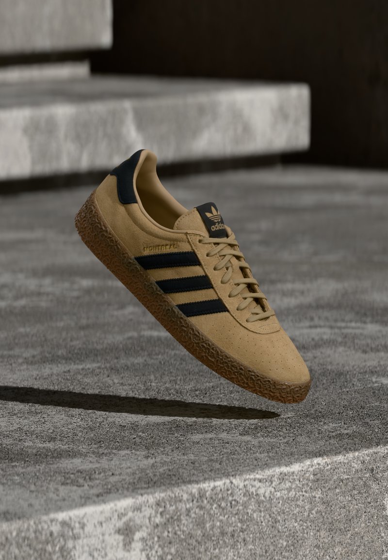 Gelber Wildledersneaker mit drei schwarzen Streifen, flachen Schnürsenkeln und strukturiertem Gummiaußensohlenprofil. Verfügt über Branding auf der Zunge und der Seite.