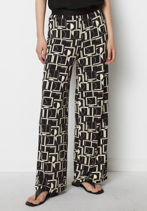 Persoon draagt zwarte, wijde broek met hoge taille en opvallende crèmekleurige geometrische vormen, zwart top en zwarte sandalen, staand binnenshuis.
