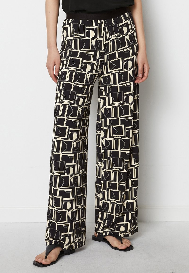 Personne portant un pantalon noir large à taille haute aux motifs géométriques crème audacieux, un haut noir et des sandales noires, debout à l'intérieur.