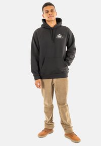 Sudadera con capucha negra y bolsillo frontal, con un logo blanco, combinada con pantalones de pana beige y zapatos marrones, de pie sobre un fondo blanco.