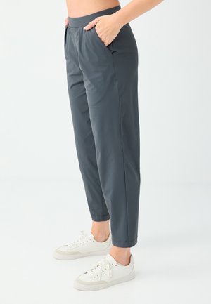 Persona che indossa pantaloni grigio scuro elasticizzati con vita affusolata e scarpe da ginnastica bianche, con le mani nelle tasche, su uno sfondo chiaro uniforme.