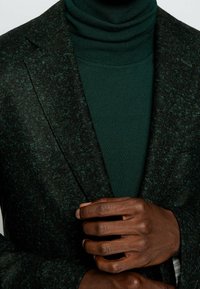 Mörkgrön texturerad blazer med slagkrage, bärs över en mörkgrön polotröja. Närbild av händer placerade nära blazern.