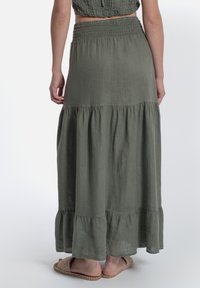 Gonna lunga maxi a balze di colore verde oliva, realizzata in tessuto leggero con vita arricciata e orlo fluttuante, abbinata a sandali piatti.