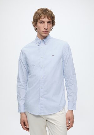 Young man with curly hair wearing a light blue striped button-up shirt and off-white pants against a plain white background.