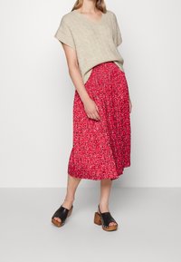 Un pull beige en maille torsadée à manches courtes associé à une jupe midi à imprimé floral rouge. Des sabots noirs à plateforme avec des semelles en bois complètent la tenue.