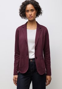 Blazer bordeaux con due tasche frontali, chiusura a un bottone e un tessuto morbido e strutturato, abbinato a una maglietta bianca e jeans scuri.