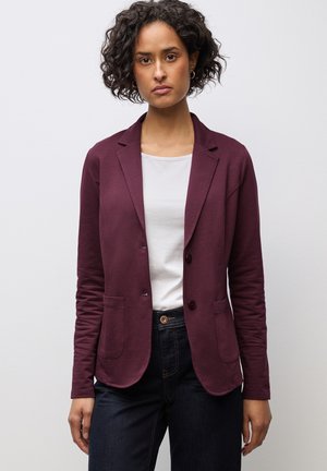 Bordeaux blazer met twee voorkezen, single-button sluiting en een zachte, gestructureerde stof, gecombineerd met een witte top en donkere jeans.
