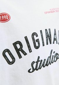 Bijela majica s crnim grafičkim tekstom "ORIGINAL studio" i crvenom oznakom "1990", od glatkog pamučnog materijala s ležernim krojem.