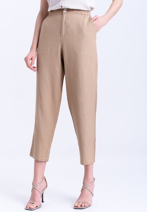 Frau trägt beige hoch taillierte, verkürzte Hose mit vorderen Knöpfen und Taschen, kombiniert mit beigen, Riemchen-Sandaletten mit hohem Absatz.