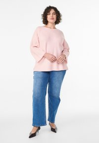 Light pink textured sweater with wide bell sleeves, paired with straight-leg blue jeans and black heeled shoes, standing against a white background.