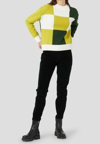 Pull en tricot avec un design coloré en vert, jaune et blanc. Manches et ourlet côtelés. Associé à un pantalon en velours côtelé noir et des bottes noires.
