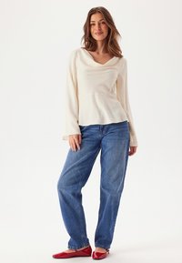 Krämfärgad långärmad blus med båtringning, kombinerad med ljusblå denimjeans och röda ballerinas. Slät textur och avslappnad passform.