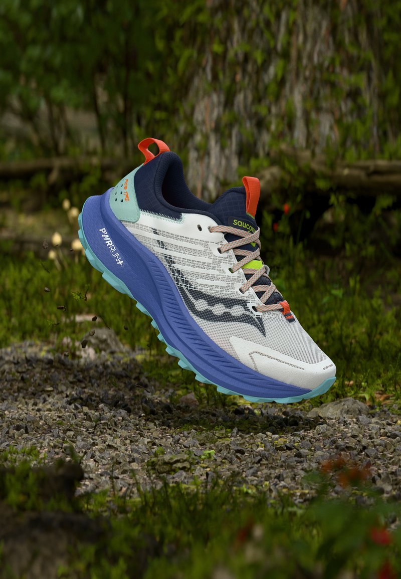 Λευκό, μπλε και πράσινο παπούτσι για τρέξιμο σε μονοπάτι με πορτοκαλί λεπτομέρειες να αιωρείται πάνω από πετρώδες δάσος κοντά σε κορμό δέντρου καλυμμένο με βρύα.