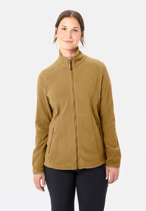 Jonge vrouw staat met het gezicht naar voren, draagt een beige fleecejack met rits en zwarte broek tegen een effen lichte achtergrond.