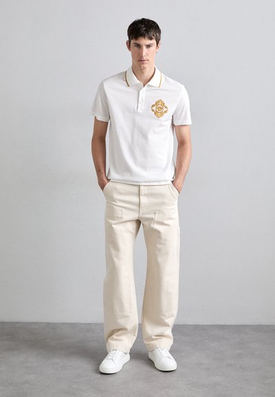 White cotton polo shirt with gold emblem detail, paired with light beige cotton trousers. Model wears white sneakers. Neutral background.