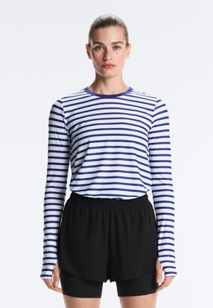 Žena čelom vpred, oblečená v bielej košeli s dlhým rukávom s námorníckymi modrými horizontálnymi pruhmi a čierne vrstvené športové šortky.