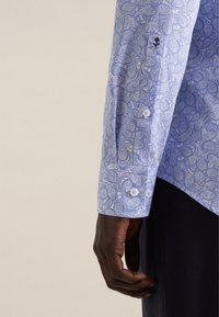 Blauw overhemd met lange mouwen, voorzien van een paisleypatroon, witte knopen en een zwart bloemenaccent op de manchetten. Glad, lichtgewicht stof.