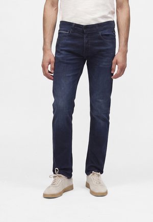 Mann trägt dunkelblaue Slim-Fit-Jeans und beige Sneaker, steht vor einem schlichten weißen Hintergrund.
