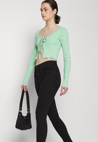 Top curto em verde menta com ribanas e mangas compridas, com um detalhe de laço na frente, combinado com jeans pretos ajustados e uma pequena mala preta.