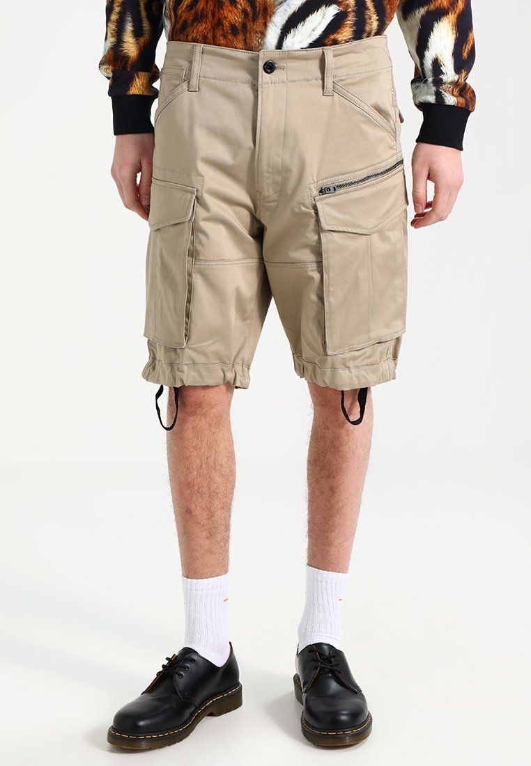 Shorts cargo beiges avec grandes poches, détail zippé et ourlets ajustables, portés avec des chaussettes blanches et des chaussures noires à lacets.