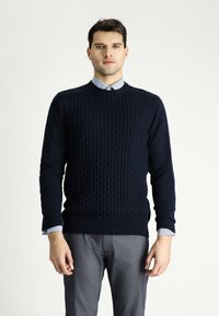 Dunkelblauer gestrickter Pullover mit strukturiertem Muster, Rundhalsausschnitt und gerippten Bündchen. Getragen über einem blau gestreiften Hemd und grauen Hosen.