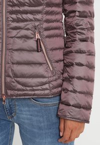 Fioletowa ocieplana kurtka o pikowanym wzorze, z zamykanymi na zamek kieszeniami i gładką, błyszczącą powierzchnią. Posiada dopasowane rękawy i różowo-złote akcesoria.