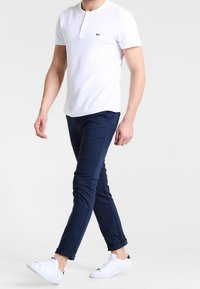 Bílá polokošile s krátkým rukávem a zeleným logem, spárovaná s tmavomodrými slim-fit kalhotami a bílými teniskami. Texturovaná tkanina, čistý design.