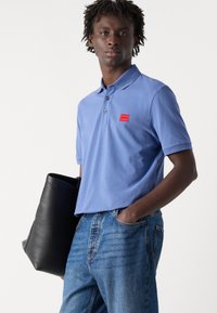 Lys blå polo skjorte med korte ermer og en rød rektangulær logo-patch, kombinert med blå denim jeans og en svart lærveske.