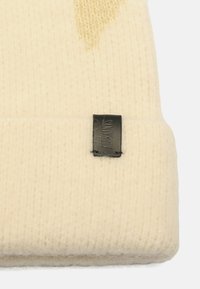 Cream-colored knit beanie with a textured pattern and a black leather label stitched on the cuff. Soft material and snug fit.
