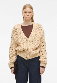 Beżowy sweter kardigan z dekoltem w serek, z dużymi teksturowanymi ściegami, dwoma dużymi guzikami i ściągaczem u dołu. Zestawiony z ciemnoniebieskimi dżinsami.