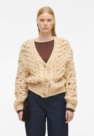 Beige Strickjacke mit V-Ausschnitt, die große strukturierte Maschen, zwei übergroße Knöpfe und einen gerippten Saum aufweist. Kombiniert mit dunklen Jeans.