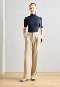 Un pull à col roulé côtelé bleu marine associé à un pantalon large plissé beige. Des chaussures à enfiler bi-couleur grises et noires complètent la tenue.