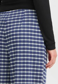 Blaue und weiße karierte Hosen aus einem gewebten Stoff, mit lockerer Passform und einer subtilen Textur. Eine Hand einer Person ruht an der Seite.