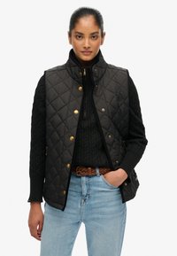 Czarna pikowana kamizelka z wysokim kołnierzem, złotymi detalami i bocznymi kieszeniami, noszona na czarnym swetrze w warkocze i niebieskich dżinsach.