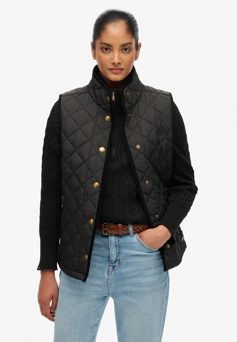 Czarna pikowana kamizelka z wysokim kołnierzem, złotymi detalami i bocznymi kieszeniami, noszona na czarnym swetrze w warkocze i niebieskich dżinsach.