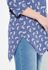 Blaue Bluse mit Muster und kurzen Ärmeln, die helle abstrakte Formen und einen abgerundeten Saum aufweist, kombiniert mit dunkelblauen Jeans.