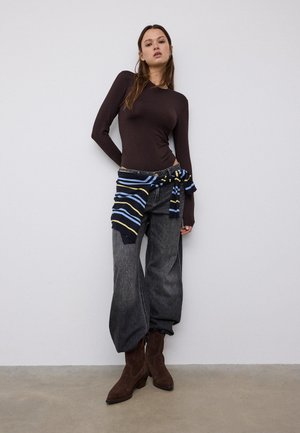 Kvinde iført en tætsiddende brun langærmet top, løse mørkegrå jeans, brune støvler og en stribet sweater bundet om taljen, stående op ad en hvid væg.