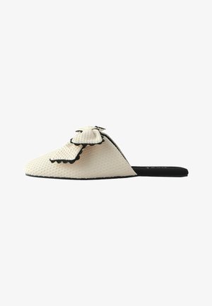 Cream textured slip-on flat with a large bow featuring black scalloped edges and a black sole.