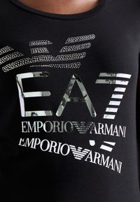 Zwarte T-shirt met een reflecterend, metallic logo ontwerp met strasstenen accenten die "EA7 Emporio Armani" spellen, gekenmerkt door verschillende tekststijlen.