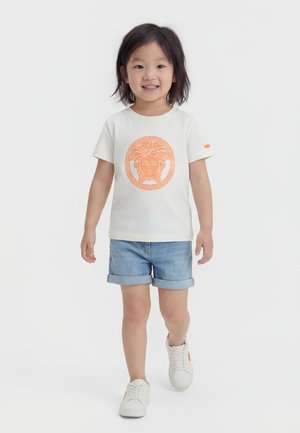 Jeune enfant marchant, portant un T-shirt blanc avec un imprimé Méduse orange, un short en jean et des baskets blanches avec des touches orange.