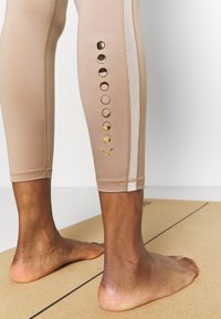 Leggings beige realizzati in materiale elasticizzato, con strisce laterali bianche e dettagli circolari dorati lungo il polpaccio esterno.