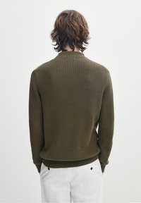 Maglione lavorato a maglia verde oliva con texture a costine, scollatura a girocollo e polsini aderenti, abbinato a pantaloni bianchi e un design minimalista.