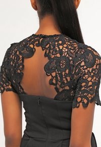 Svart klänning med genomskinlig rygg och intrikata svarta spetsdetaljer, korta ärmar och en figurnära kropp med dragkedjestängning.