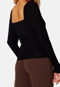 Top lungo a maniche nere a coste con scollatura quadrata e dettagli arricciati sulle spalle, abbinato a pantaloni aderenti marroni.