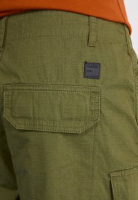 Short cargo vert avec poche à rabat et patch noir Alpha Industries porté avec une chemise orange.
