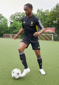 Camiseta negra de la Juventus con acentos amarillos, pantalones cortos negros y calcetas de fútbol negras con rayas blancas. Botines blancos y un balón de fútbol colorido.